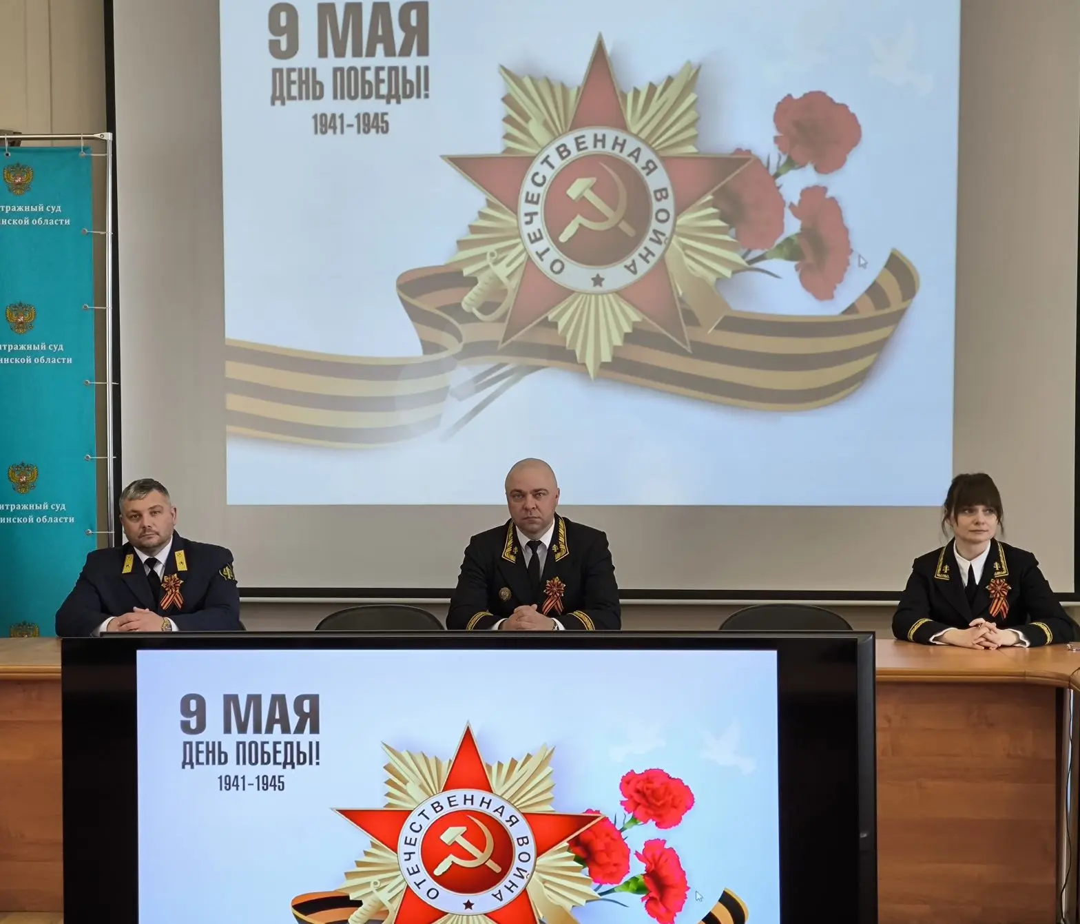 На фото изображен председатель Арбитражного суда Сахалинской области, слева администратор суда, справа заместитель председателя суда. Сзади на экране изображение 5 конечной звезды с серпом и молотом и надпись 9 Мая день Победы 1941-1945